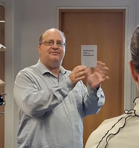 Vaaleansiniseen paitaan pukeutunut mies käyttää käsieleitä huoneessa. Nainen seisoo etualalla. Ovi ja hyllyt taustalla.