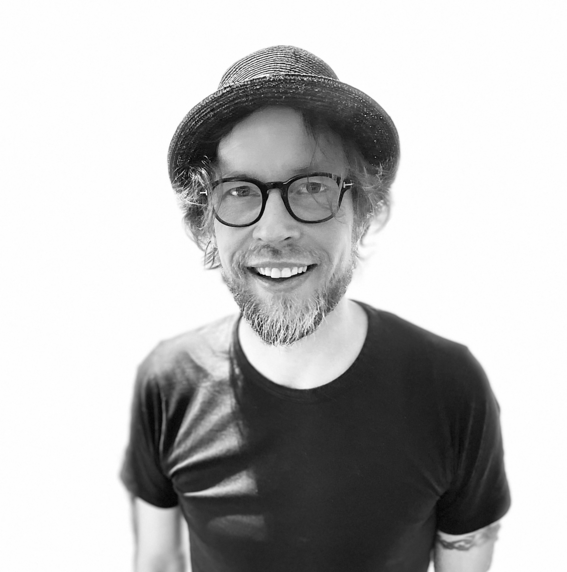 A man wearing a black T-shirt and a hat, with an arm tattoo, shown against a plain white background.