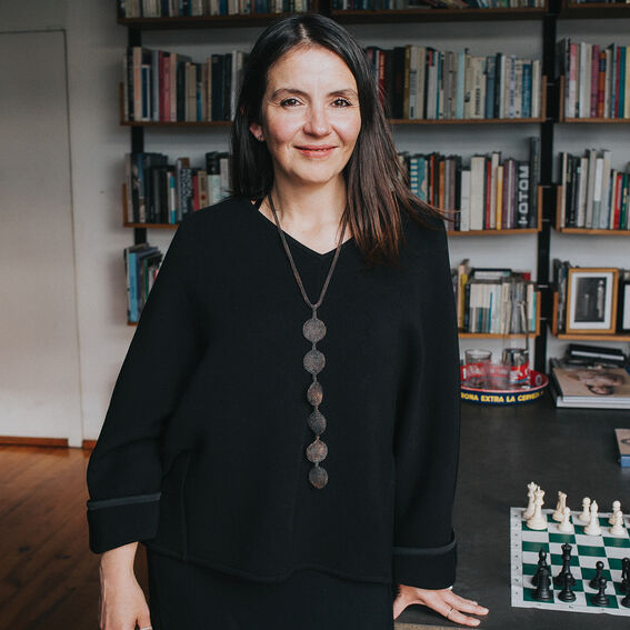 A person wearing a black top and necklace stands in a room with bookshelves and a chessboard on a table.