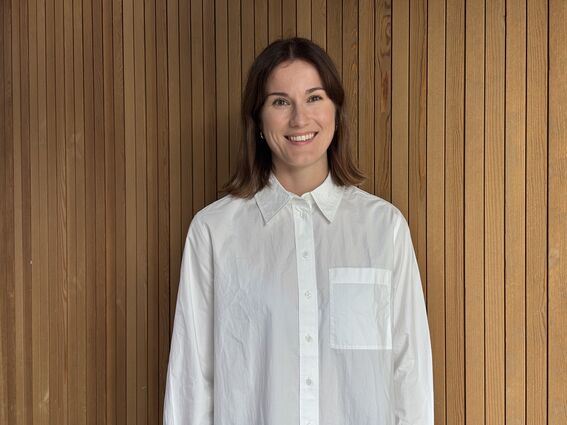 Iiris Hörhammer, joka on pukeutunut valkoiseen nappipaitaan, seisoo puurimaseinän edessä.