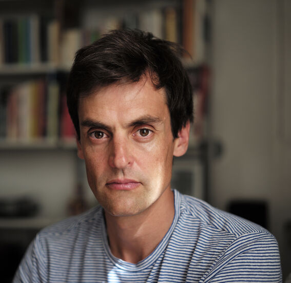 Person with dark hair wearing a blue and white striped shirt, sitting indoors with blurred bookshelves in the background.