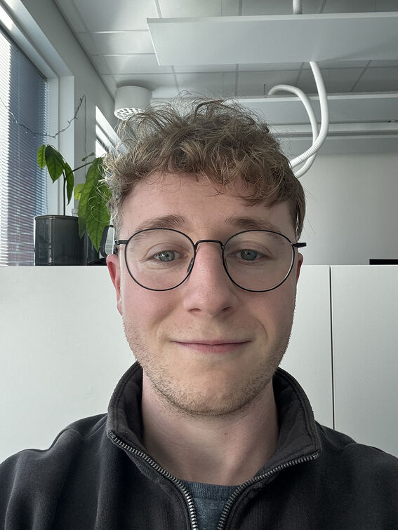 A person with curly hair is inside an office with a plant on the windowsill and white cabinetry behind them.
