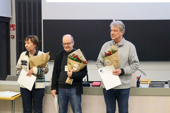 Three people holding bouquets and certificates stand in a classroom with a blackboard behind them.
