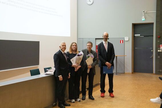 Neljä ihmistä seisoo luentosalissa pitäen kukkia ja todistuksia. Seinällä näkyy kello.