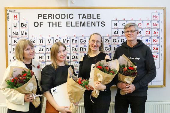 Four people holding bouquets stand in front of a periodic table poster.