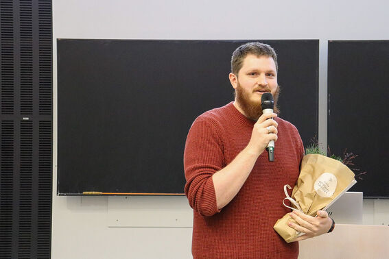 A person in a red jumper holds a microphone and a bouquet wrapped in brown paper, standing in front of a blackboard.