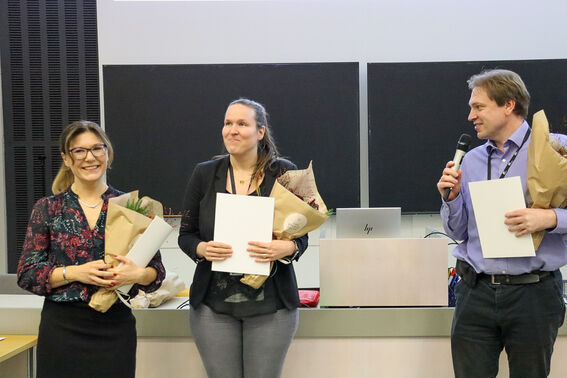 Three people holding flowers and certificates stand in front of a podium. One person is speaking into a microphone.