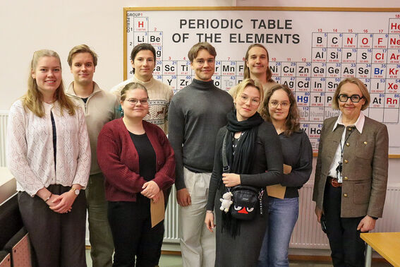 A group of people standing in front of a periodic table of elements poster in a classroom.
