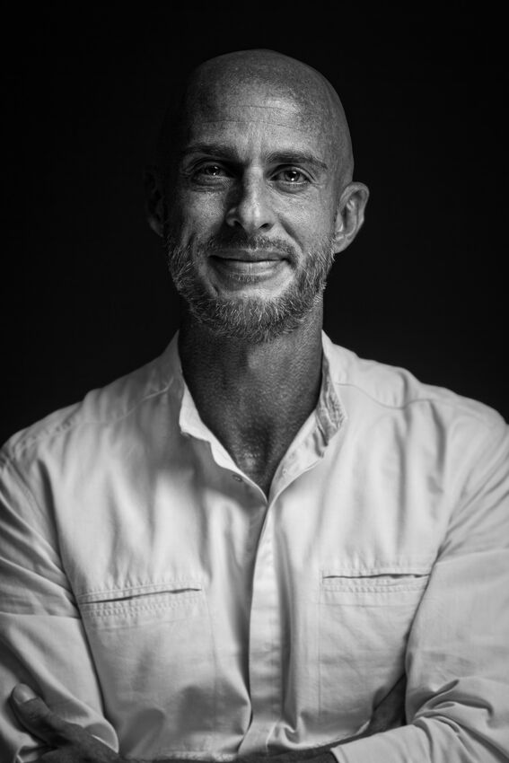 Mustavalkoinen kuva henkilöstä, joka käyttää vaaleaa paitaa ja pitää kädet ristissä tummalla taustalla.