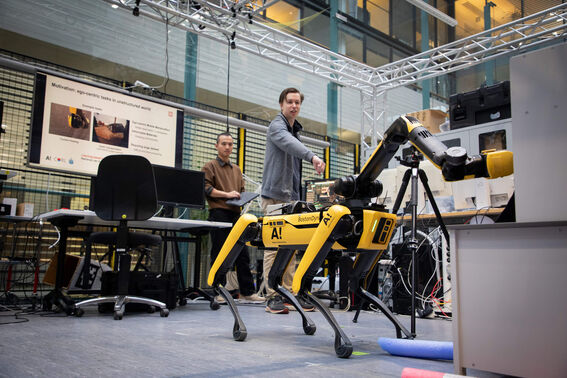 Två personer interagerar med en gul Boston Dynamics robot hund i ett tekniskt labb med skärmar och utrustning.