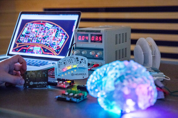 Työpöytä, jossa on elektroniikkakomponentteja, kannettava tietokone, joka näyttää piirikaavion, ja hehkuva aivomalli.