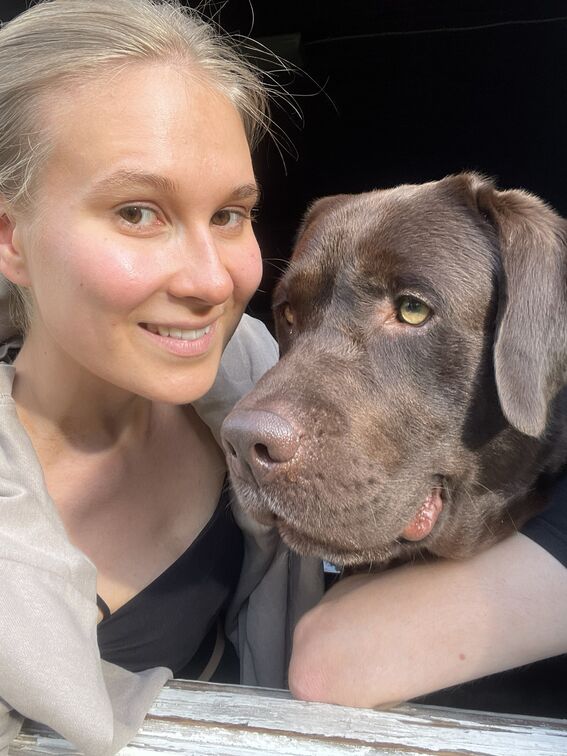 Vaaleahiuksinen henkilö mustassa topissa ja beigessä huivissa ruskean koiran vieressä. 