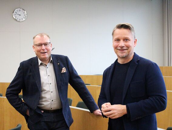 Kaksi miestä puvuissa seisoo huoneessa, jossa on puisia pöytiä ja seinäkello.