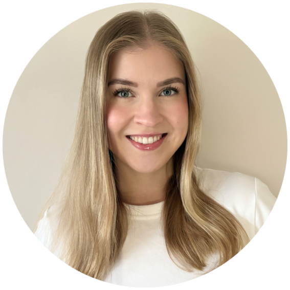 A person with long, straight blonde hair wearing a white shirt, standing against a light-coloured wall.
