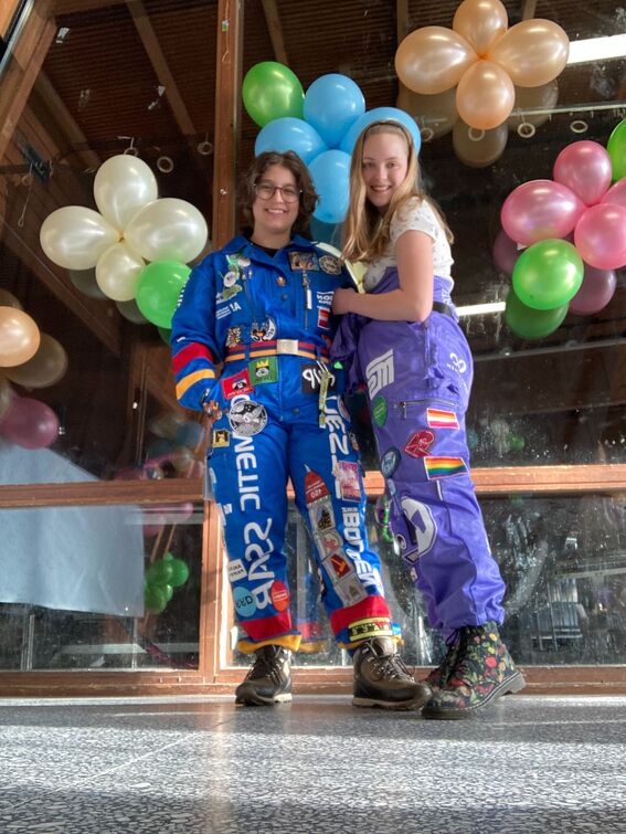 Two people in colourful student overalls stand in front of a window decorated with balloons.