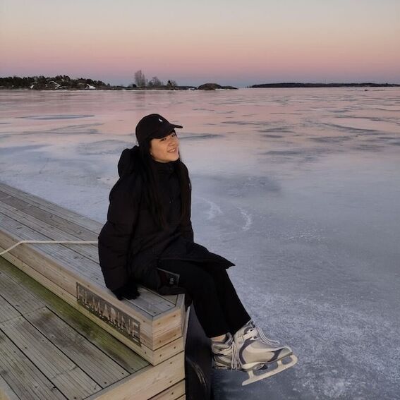 A person in winter clothing sits on a wooden dock by a frozen lake at sunset. Ice skates are worn.