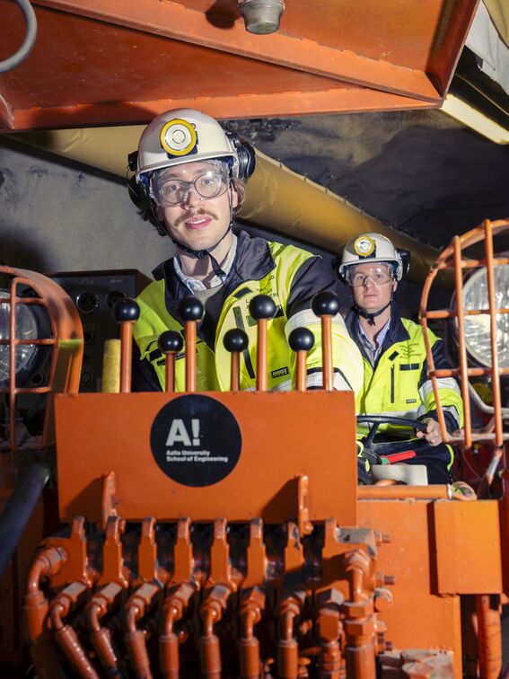 Two workers in high-visibility jackets and helmets operate machinery in an industrial setting.