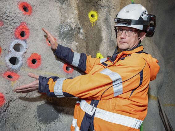 A person in an orange safety jacket gestures towards a rock wall with coloured drill holes.