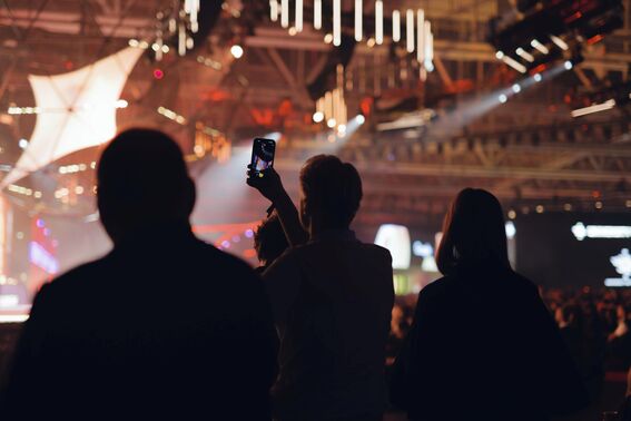 Three people in silhouette at an indoor event. One person is holding up a phone to take a photo. Bright lights in the background.