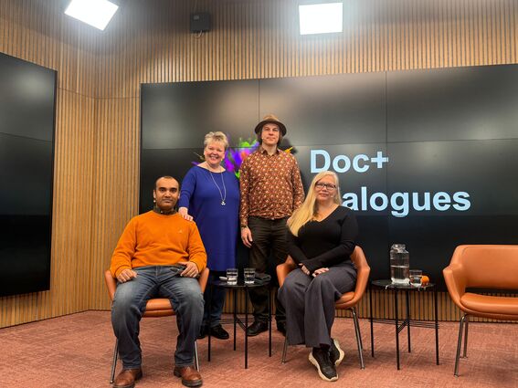 Neljä ihmistä istuu ja seisoo huoneessa, jossa on puuseinät ja näyttö, jossa lukee 'Doc+ Dialogues'.