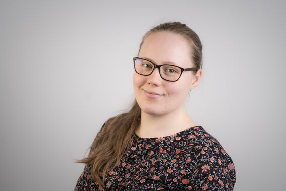 Karola Salminen, jolla on pitkät ruskeat hiukset ja musta kukkakuvioinen paita harmaata taustaa vasten.