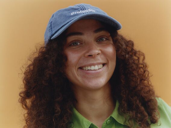 Person with curly hair wearing a blue cap and green shirt, standing against a beige background.