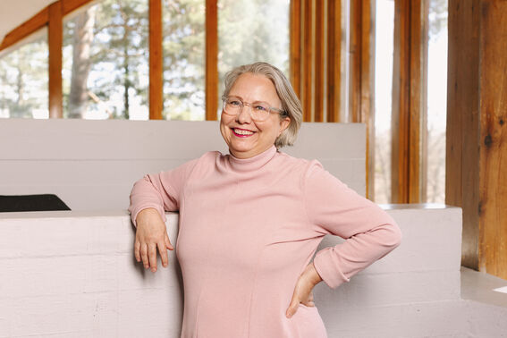 A person in a pink jumper leaning on a white wall. The background shows wooden pillars and large windows with trees outside.
