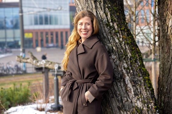 Person with long hair leaning against a tree wearing a brown coat. Blurred urban background.