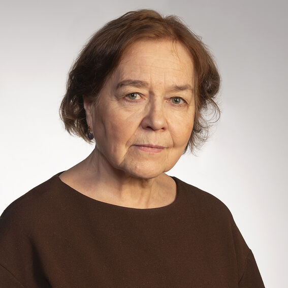 Iäkkäämpi nainen, ruskeat hiukset ja tummanruskea paita vaaleaa taustaa vasten