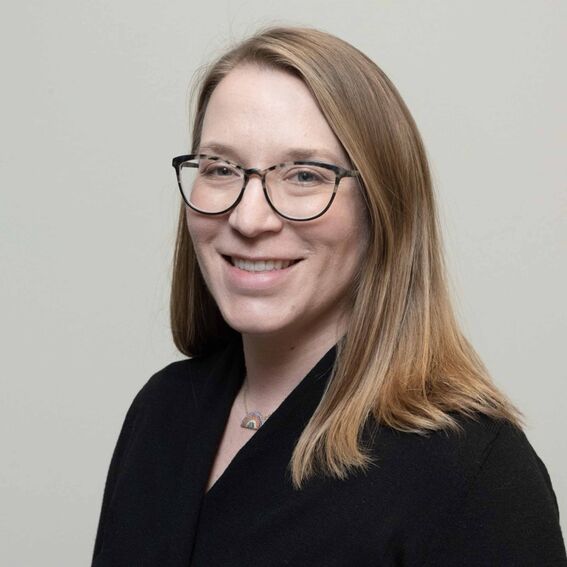 Henkilö, olkapäille ulottuvat vaaleanruskeat hiukset, musta paita, sateenkaarikaulakoru, harmaa tausta, kasvot blurattu