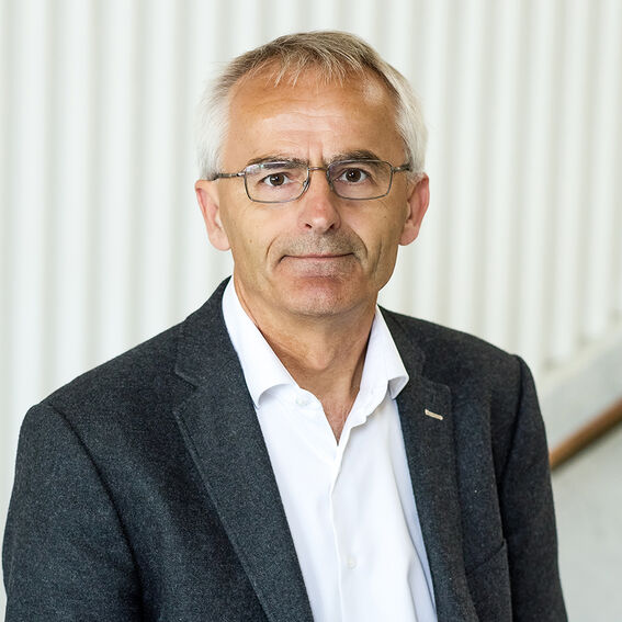 Iäkkäämpi mies tummassa bleiserissä ja valkoisessa paidassa, vaalea raidallinen tausta