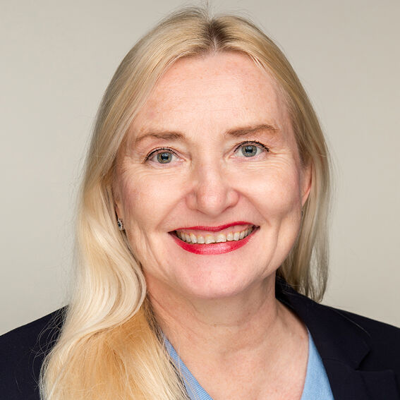 Pitkähiuksinen vaalea aikuinen tummassa jakussa ja vaaleansinisessä paidassa, neutraali tausta