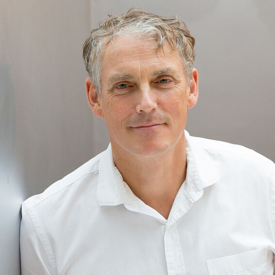 Aikuinen, lyhyet harmahtavat hiukset, valkoinen paita, vaaleanharmaa seinä taustalla