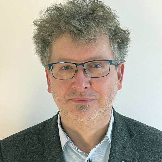 Henkilö, jolla lyhyet kiharat harmaat hiukset, puku ja vaalea paita, vaaleaa seinää vasten