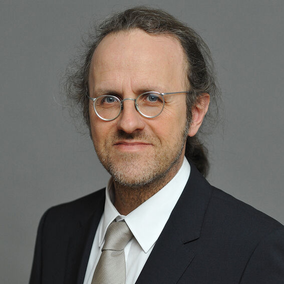 Henkilö tummassa puvussa, valkoinen paita ja vaalea solmio, harmaata taustaa vasten