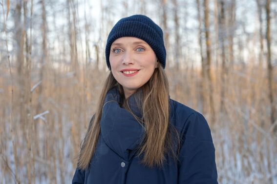 Person wearing a dark blue jacket and knit hat, standing outside with dry, tall grass and trees in the background.