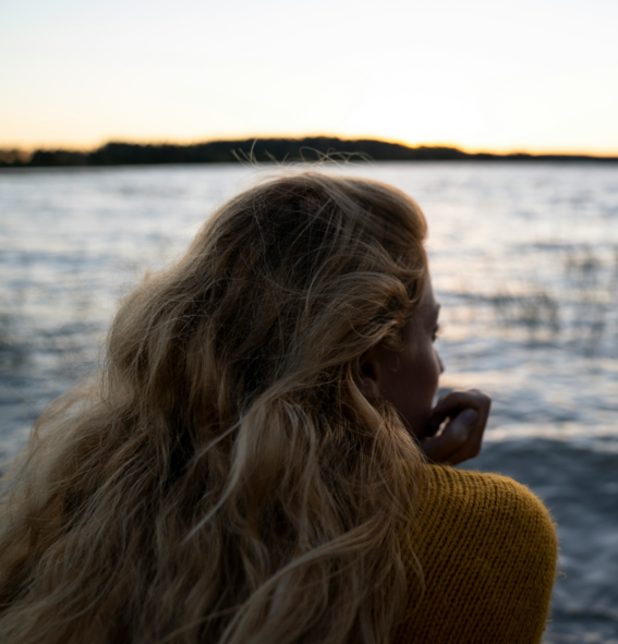 A person with long wavy hair stands near a calm lake at sunset, wearing a mustard yellow sweater. Image by Hendrik morkel