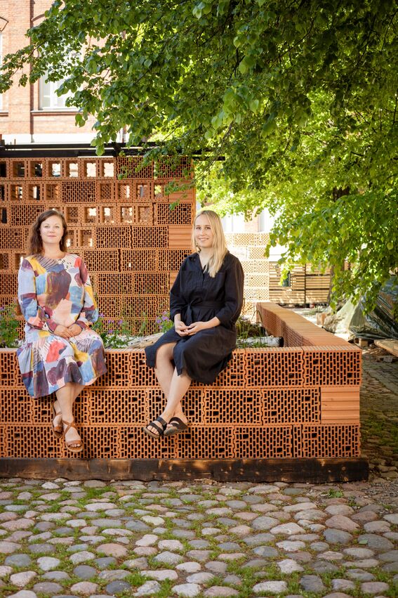 Kaksi naista istuu matalalla tiiliseinällä pihan puun alla, juttelee ja rentoutuu ulkona.