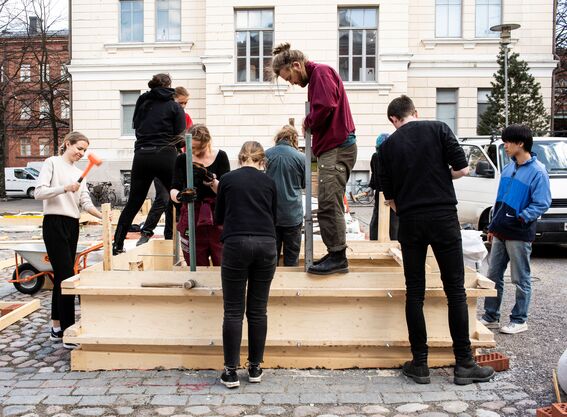 Ryhmä ihmisiä rakentaa ulkona puista tasoa mukulakivillä vaalean rakennuksen edessä