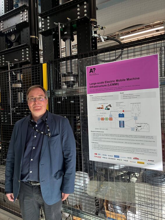 Person in blue blazer stands by industrial hydraulics machinery and a large pink research poster behind wire fencing.