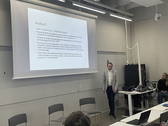 Person giving a lecture on artefacts, standing by a projected slide in a modern grey classroom.