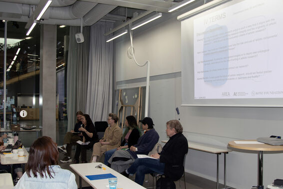 People sit in a modern classroom, discussing while a large presentation slide is projected on a white wall.
