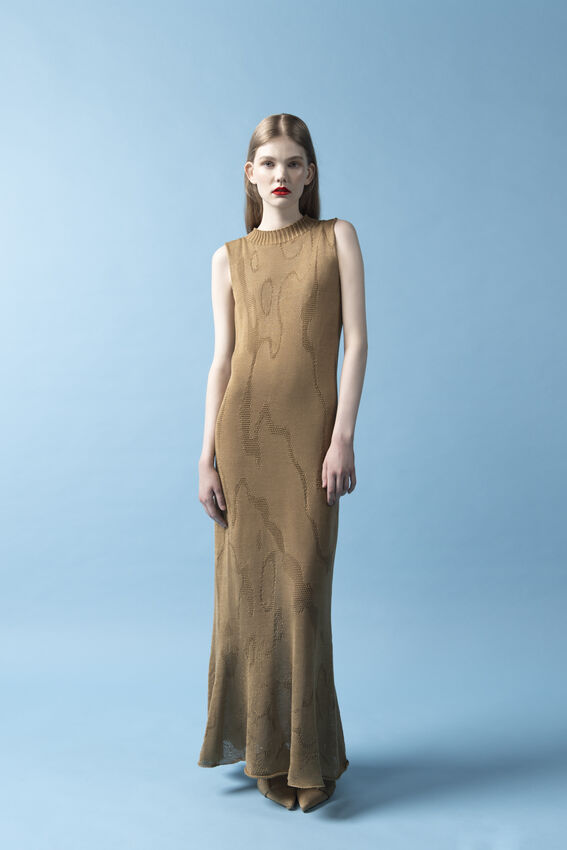 Person in a long sleeveless tan knit dress with wavy pattern, standing against a plain light blue backdrop.