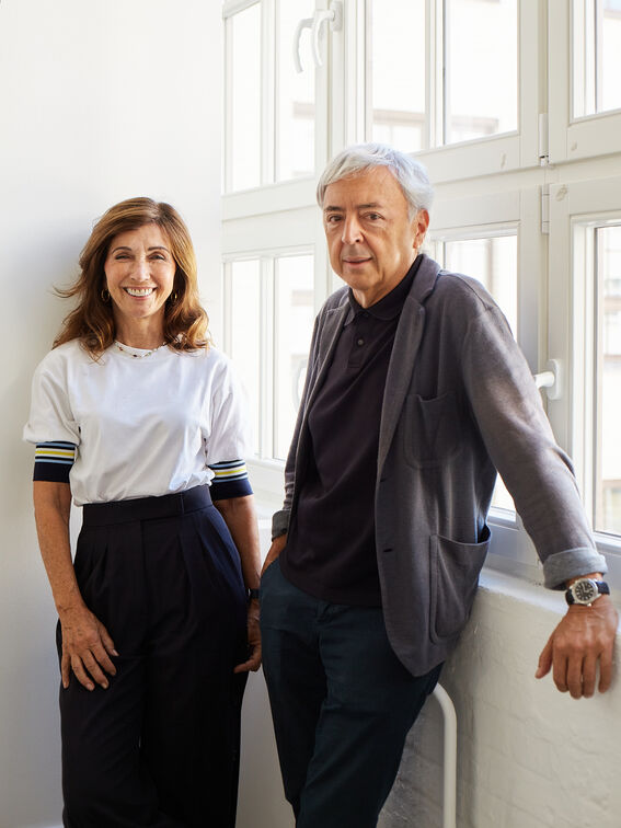 Two adults with blurred faces stand by tall windows, casually dressed in dark trousers and light tops