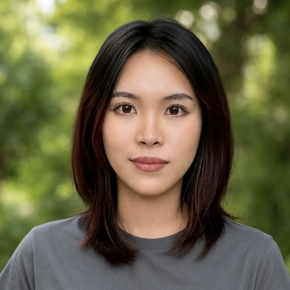 Person with shoulder-length dark hair in grey T-shirt, blurred face, standing outdoors with green trees behind