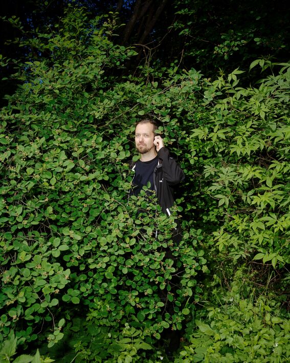Person in dark clothes standing partly hidden among dense green bushes in a forest