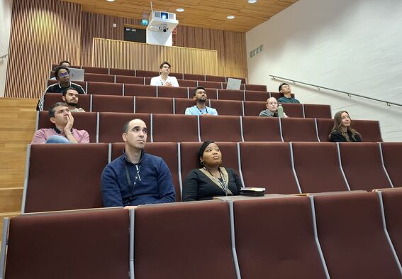 Small group of students sit spaced out in a brown lecture hall with laptops and notebooks