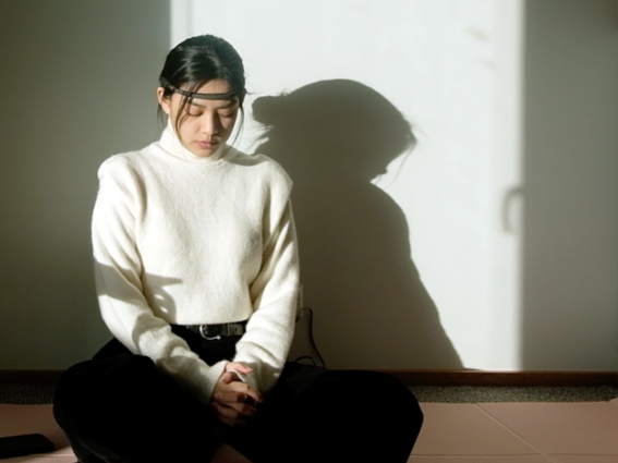 Person in white jumper sitting cross-legged on floor, face blurred, shadow cast on light wall