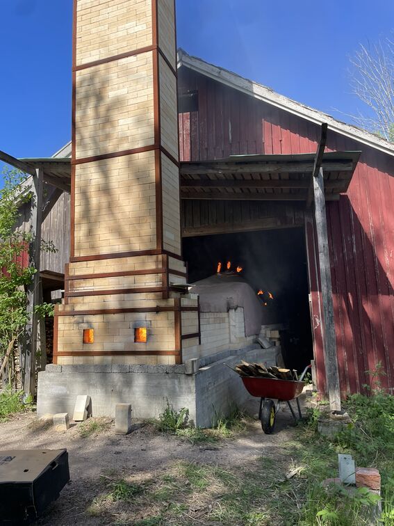 Tall brick kiln with glowing fireboxes beside a red wooden barn, wheelbarrow of wood in front