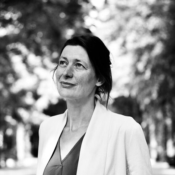 Black-and-white portrait of a woman outdoors, face blurred, wearing a light jacket over a dark top